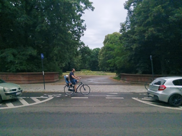 A man is riding a bicycle on a paved road with a child seated behind him, near a park area surrounded by lush green trees. There are two cars parked on either side of the road, and the atmosphere is calm and overcast.