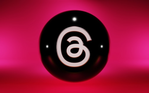 A black sphere with a stylized white 'a' symbol, set against a vibrant pink background. The sphere is glossy, reflecting light that creates small highlights on its surface.