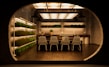 A modern, indoor garden kitchen with shelves of green plants on the left and background walls. The central focus is a kitchen island with a dark countertop, adorned with small ornamental plants and a set of matching four white wireframe chairs. The ceiling features a series of large, rectangular, warm-lit fixtures, creating a cozy ambiance and reflecting against the floor tiles.