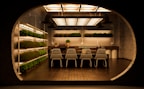 A modern, indoor garden kitchen with shelves of green plants on the left and background walls. The central focus is a kitchen island with a dark countertop, adorned with small ornamental plants and a set of matching four white wireframe chairs. The ceiling features a series of large, rectangular, warm-lit fixtures, creating a cozy ambiance and reflecting against the floor tiles.