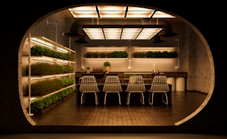 A modern, indoor garden kitchen with shelves of green plants on the left and background walls. The central focus is a kitchen island with a dark countertop, adorned with small ornamental plants and a set of matching four white wireframe chairs. The ceiling features a series of large, rectangular, warm-lit fixtures, creating a cozy ambiance and reflecting against the floor tiles.