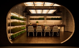 A modern, indoor garden kitchen with shelves of green plants on the left and background walls. The central focus is a kitchen island with a dark countertop, adorned with small ornamental plants and a set of matching four white wireframe chairs. The ceiling features a series of large, rectangular, warm-lit fixtures, creating a cozy ambiance and reflecting against the floor tiles.