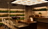 A modern kitchen with a sleek design featuring marble countertops and a contemporary lighting fixture. Rows of small potted plants line the shelves on the walls, adding greenery to the space. There are white bar stools with metal frames positioned along a counter. Decorative bottles and vases with plants are placed on the countertops, contributing to a warm and inviting atmosphere.