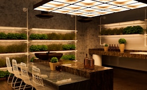 A modern kitchen with a sleek design featuring marble countertops and a contemporary lighting fixture. Rows of small potted plants line the shelves on the walls, adding greenery to the space. There are white bar stools with metal frames positioned along a counter. Decorative bottles and vases with plants are placed on the countertops, contributing to a warm and inviting atmosphere.