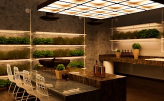 A modern kitchen with a sleek design featuring marble countertops and a contemporary lighting fixture. Rows of small potted plants line the shelves on the walls, adding greenery to the space. There are white bar stools with metal frames positioned along a counter. Decorative bottles and vases with plants are placed on the countertops, contributing to a warm and inviting atmosphere.