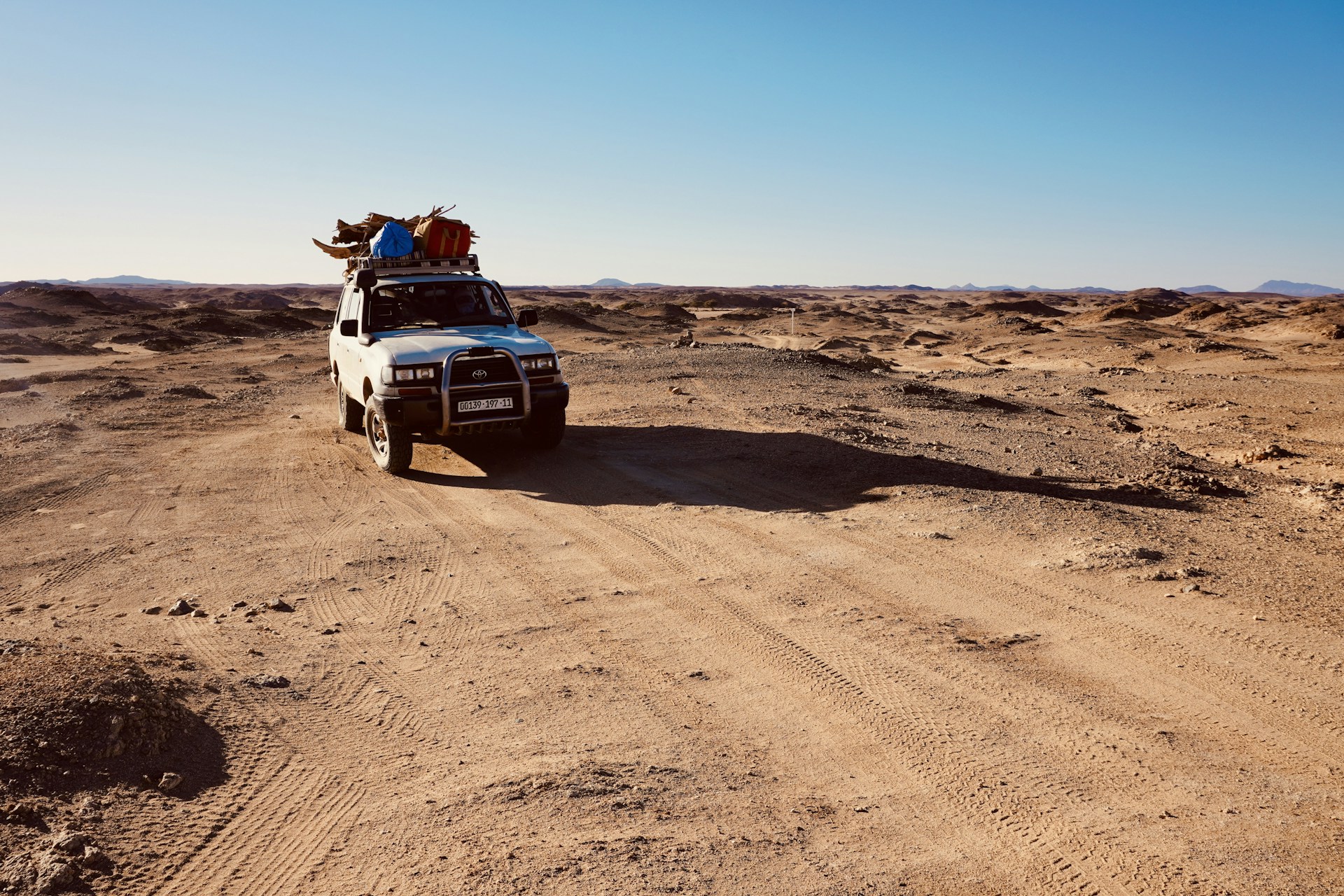 A white off-road vehicle travels through a barren desert landscape under a clear blue sky. The vehicle is equipped with various items secured on its roof, including colorful bags and supplies. The surrounding terrain is rugged and arid, with mountains visible in the distant horizon. The sunlight casts long shadows across the sandy ground.