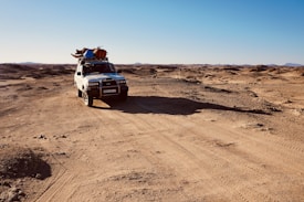 A white off-road vehicle travels through a barren desert landscape under a clear blue sky. The vehicle is equipped with various items secured on its roof, including colorful bags and supplies. The surrounding terrain is rugged and arid, with mountains visible in the distant horizon. The sunlight casts long shadows across the sandy ground.