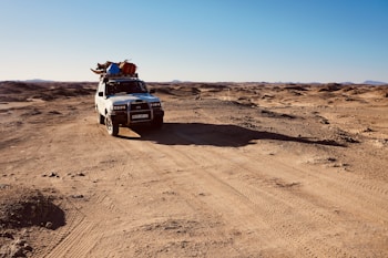 A white off-road vehicle travels through a barren desert landscape under a clear blue sky. The vehicle is equipped with various items secured on its roof, including colorful bags and supplies. The surrounding terrain is rugged and arid, with mountains visible in the distant horizon. The sunlight casts long shadows across the sandy ground.