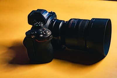 A digital camera with a large lens, resting on a smooth, yellow surface. The camera is black with noticeable control dials on the top and branding on the side of the lens.