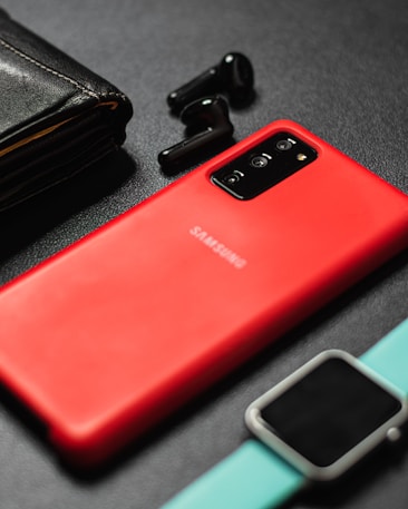 A vibrant red Samsung smartphone is placed on a dark textured surface, accompanied by a black leather wallet, a pair of black wireless earbuds, and a smartwatch with a turquoise band. The layout appears organized, showcasing a selection of modern electronic devices and personal accessories.