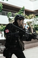 A person dressed in tactical gear and a gas mask is holding a large firearm. The scene includes some vegetation and appears to be set in an urban environment with modern architecture. The person's uniform is predominantly black with a red and white emblem on the sleeve.