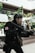A person dressed in tactical gear and a gas mask is holding a large firearm. The scene includes some vegetation and appears to be set in an urban environment with modern architecture. The person's uniform is predominantly black with a red and white emblem on the sleeve.