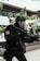 A person dressed in tactical gear and a gas mask is holding a large firearm. The scene includes some vegetation and appears to be set in an urban environment with modern architecture. The person's uniform is predominantly black with a red and white emblem on the sleeve.
