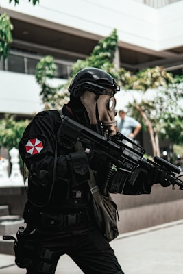 A person dressed in tactical gear and a gas mask is holding a large firearm. The scene includes some vegetation and appears to be set in an urban environment with modern architecture. The person's uniform is predominantly black with a red and white emblem on the sleeve.