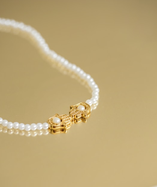 A pearl necklace with a gold clasp featuring a pair of ornate designs, each adorned with a central pearl, is laid elegantly on a golden surface.