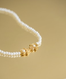 A pearl necklace with a gold clasp featuring a pair of ornate designs, each adorned with a central pearl, is laid elegantly on a golden surface.