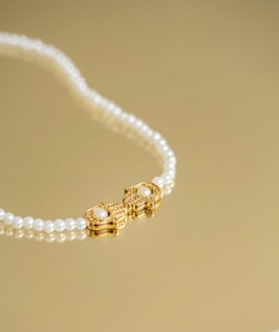 A pearl necklace with a gold clasp featuring a pair of ornate designs, each adorned with a central pearl, is laid elegantly on a golden surface.