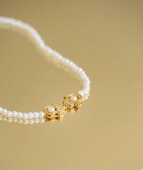 A pearl necklace with a gold clasp featuring a pair of ornate designs, each adorned with a central pearl, is laid elegantly on a golden surface.