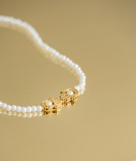 A pearl necklace with a gold clasp featuring a pair of ornate designs, each adorned with a central pearl, is laid elegantly on a golden surface.