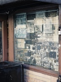An assortment of locks and keys is displayed in a shop window. The window is framed with brown wood, and a wire mesh covers the glass. Various types of locks, including padlocks and antique keys, are visible. There are also licenses and signs visible inside the shop.