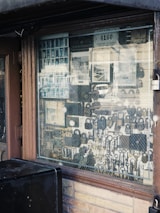 An assortment of locks and keys is displayed in a shop window. The window is framed with brown wood, and a wire mesh covers the glass. Various types of locks, including padlocks and antique keys, are visible. There are also licenses and signs visible inside the shop.