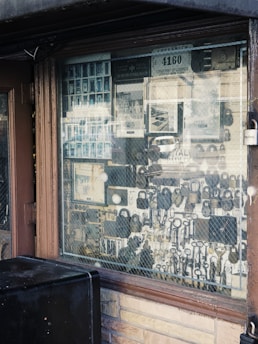 An assortment of locks and keys is displayed in a shop window. The window is framed with brown wood, and a wire mesh covers the glass. Various types of locks, including padlocks and antique keys, are visible. There are also licenses and signs visible inside the shop.
