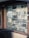 An assortment of locks and keys is displayed in a shop window. The window is framed with brown wood, and a wire mesh covers the glass. Various types of locks, including padlocks and antique keys, are visible. There are also licenses and signs visible inside the shop.