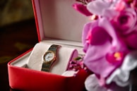 A luxury watch is displayed in a red and white box, accompanied by vibrant purple and pink flowers. The watch has a metal strap and a classic round face, suggesting elegance and sophistication.