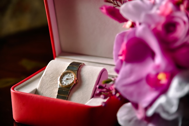 A luxury watch is displayed in a red and white box, accompanied by vibrant purple and pink flowers. The watch has a metal strap and a classic round face, suggesting elegance and sophistication.