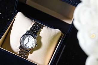 A silver wristwatch with a white dial and Roman numeral hour markers is displayed in an open black box lined with beige fabric. The watch is positioned on a cushioned pad inside the box. To the right, an out-of-focus white object, possibly a flower, is partially visible.