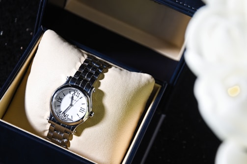 A silver wristwatch with a white dial and Roman numeral hour markers is displayed in an open black box lined with beige fabric. The watch is positioned on a cushioned pad inside the box. To the right, an out-of-focus white object, possibly a flower, is partially visible.