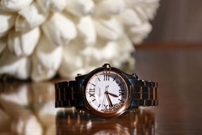A sophisticated wristwatch with a gold and black metal band is positioned prominently in front of a blurred background. The watch face is white with Roman numerals and has a few small decorative stones at the bottom. The reflective surface underneath enhances the elegance of the watch. Behind it, there are soft white petals, possibly flowers or a decorative arrangement.
