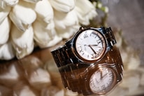 A luxury wristwatch with a metal bracelet and a white dial featuring Roman numerals is elegantly displayed on a reflective surface. The watch is accompanied by an arrangement of white flowers, adding a touch of sophistication.