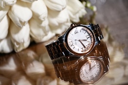 A luxury wristwatch with a metal bracelet and a white dial featuring Roman numerals is elegantly displayed on a reflective surface. The watch is accompanied by an arrangement of white flowers, adding a touch of sophistication.