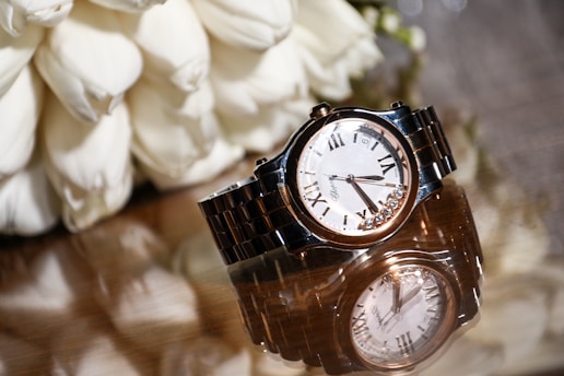 A luxury wristwatch with a metal bracelet and a white dial featuring Roman numerals is elegantly displayed on a reflective surface. The watch is accompanied by an arrangement of white flowers, adding a touch of sophistication.