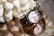 A luxury wristwatch with a metal bracelet and a white dial featuring Roman numerals is elegantly displayed on a reflective surface. The watch is accompanied by an arrangement of white flowers, adding a touch of sophistication.
