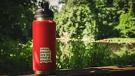 A red water bottle is placed on a wooden ledge with a forest and a body of water in the background. The bottle has a black lid and a sticker that reads 'camp people are my kind of people'. The setting suggests an outdoor, nature-focused environment with lush greenery.