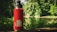 A red water bottle is placed on a wooden ledge with a forest and a body of water in the background. The bottle has a black lid and a sticker that reads 'camp people are my kind of people'. The setting suggests an outdoor, nature-focused environment with lush greenery.