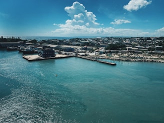 A coastal town with several buildings along the waterfront and a prominent pier extending into the calm, turquoise sea. The sky is clear with a few scattered clouds, creating a serene and relaxed atmosphere.