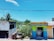 A bright and colorful street scene featuring a small commercial building with a painted facade. The left side shows an older building with faded paint and a palm tree in the background. On the right, a vividly painted shop with the sign 'BANCA MJP', offering fast and secure payment services, is visible. The setting is tropical, with lush greenery and a clear blue sky overhead.