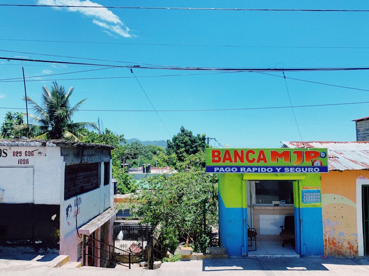 A bright and colorful street scene featuring a small commercial building with a painted facade. The left side shows an older building with faded paint and a palm tree in the background. On the right, a vividly painted shop with the sign 'BANCA MJP', offering fast and secure payment services, is visible. The setting is tropical, with lush greenery and a clear blue sky overhead.
