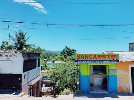 A bright and colorful street scene featuring a small commercial building with a painted facade. The left side shows an older building with faded paint and a palm tree in the background. On the right, a vividly painted shop with the sign 'BANCA MJP', offering fast and secure payment services, is visible. The setting is tropical, with lush greenery and a clear blue sky overhead.
