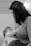 A black-and-white photograph of a woman breastfeeding a baby. The woman has dark hair and is wearing a long-sleeved shirt. The baby, wearing a patterned outfit, is cradled in her arms. The background is softly blurred, with a hint of framed or patterned elements on the wall.