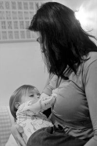 A black-and-white photograph of a woman breastfeeding a baby. The woman has dark hair and is wearing a long-sleeved shirt. The baby, wearing a patterned outfit, is cradled in her arms. The background is softly blurred, with a hint of framed or patterned elements on the wall.