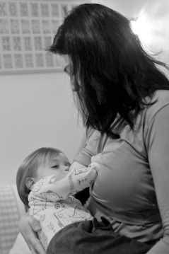 A black-and-white photograph of a woman breastfeeding a baby. The woman has dark hair and is wearing a long-sleeved shirt. The baby, wearing a patterned outfit, is cradled in her arms. The background is softly blurred, with a hint of framed or patterned elements on the wall.