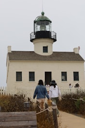A lighthouse with a white facade and green-topped lantern room, surrounded by a small picket fence. Several people are walking towards or standing near the entrance. The foreground features dry vegetation and a wooden pathway leading to the structure.