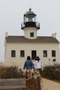 A lighthouse with a white facade and green-topped lantern room, surrounded by a small picket fence. Several people are walking towards or standing near the entrance. The foreground features dry vegetation and a wooden pathway leading to the structure.