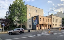 A street view featuring a three-story building with a mural on one of its walls. The building appears to be an institutional or public structure, with a sign reading 'XXILO' and additional text below it. There is a metal fence along the perimeter of the building. Trees, both green and maroon, are visible around the building. A car is parked on the street in front of the building. Several road signs and a streetlight are also visible.