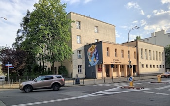 A street view featuring a three-story building with a mural on one of its walls. The building appears to be an institutional or public structure, with a sign reading 'XXILO' and additional text below it. There is a metal fence along the perimeter of the building. Trees, both green and maroon, are visible around the building. A car is parked on the street in front of the building. Several road signs and a streetlight are also visible.