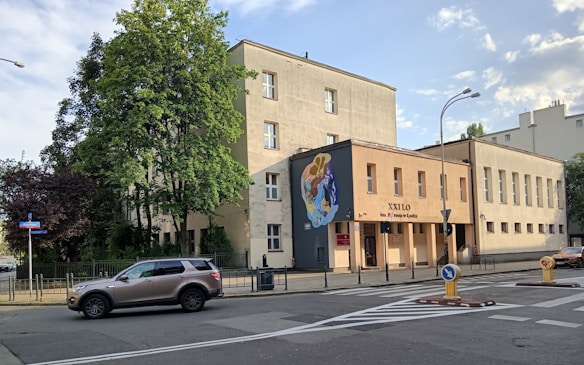 A street view featuring a three-story building with a mural on one of its walls. The building appears to be an institutional or public structure, with a sign reading 'XXILO' and additional text below it. There is a metal fence along the perimeter of the building. Trees, both green and maroon, are visible around the building. A car is parked on the street in front of the building. Several road signs and a streetlight are also visible.
